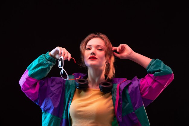 Een vooraanzicht moderne jonge dame in kleurrijke jas oranje t-shirt met zwarte koptelefoon en zonnebril poseren op de zwarte achtergrond dansen moderne mode