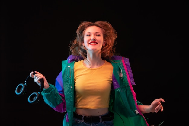 Een vooraanzicht moderne jonge dame in kleurrijke jas oranje t-shirt met zwarte koptelefoon poseren gelukkig op de zwarte achtergrond dansen moderne mode