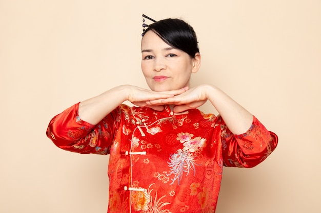 Een vooraanzicht mooie Japanse geisha in traditionele rode Japanse jurk met haar stokken poseren met haar handen staande op de crème achtergrond ceremonie onderhoudend oosten van Japan