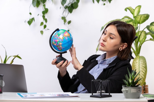 Een vooraanzicht mooie jonge zakenvrouw in zwarte jas en blauw shirt observeren kleine wereldbol voor tafel zakelijke baan kantoor