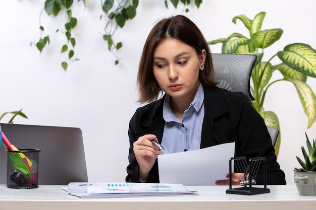 Een vooraanzicht mooie jonge zakenvrouw in zwarte jas en blauw shirt werken met laptop en documenten voor tafel zakelijke baan kantoor