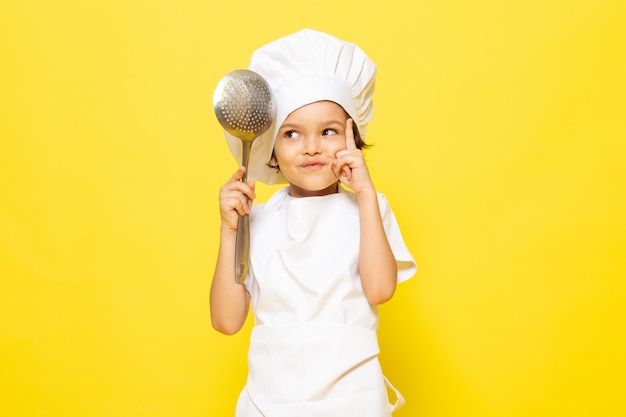 Een vooraanzicht schattig klein kind in witte kok pak en witte kok GLB met grote lepel op de gele muur kind koken keuken eten