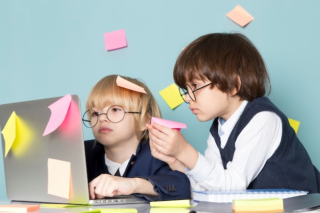 Een vooraanzicht schattige zakelijke jongen in blauwe klassieke pak poseren voor zilveren laptop samen met andere jongen bespreken lijm notities werken zakelijke werkmode