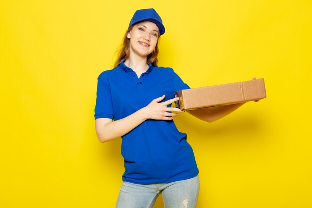 Een vooraanzicht vrouwelijke aantrekkelijke koerier in blauw poloshirt blauw GLB en jeans die pakket houden glimlachend op de gele baan van de achtergrondvoedseldienst