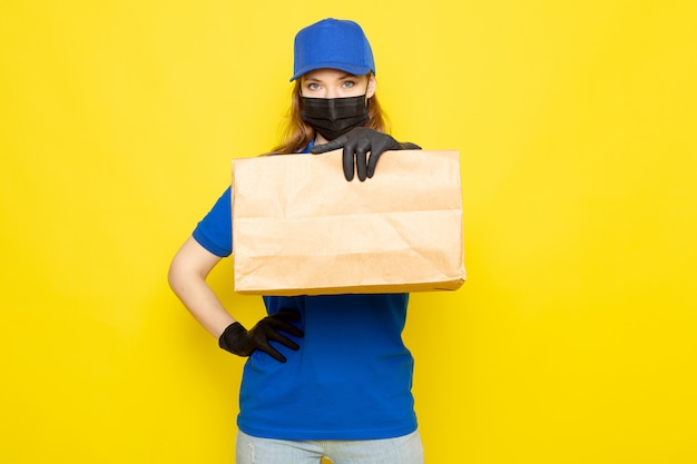 Een vooraanzicht vrouwelijke aantrekkelijke koerier in blauw poloshirt blauwe pet en jeans met pakket in zwarte handschoenen zwart beschermend masker op de gele achtergrond food service baan