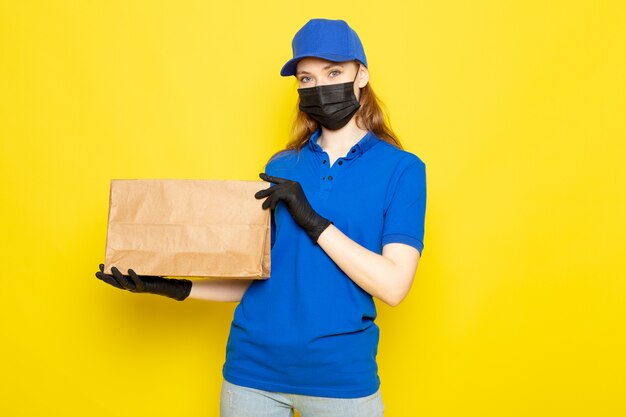 Een vooraanzicht vrouwelijke aantrekkelijke koerier in blauw poloshirt blauwe pet en jeans met pakket in zwarte handschoenen zwart beschermend masker op de gele achtergrond food service baan