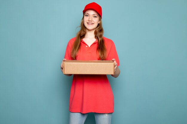 Een vooraanzicht vrouwelijke aantrekkelijke koerier in rood poloshirt rood GLB en jeans die pakket het stellen houden glimlachend op de blauwe baan van de achtergrondvoedseldienst