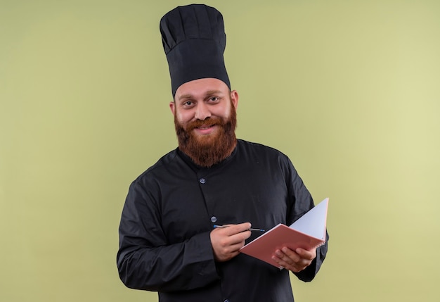 Een vrolijke bebaarde chef-kokmens in zwart uniform die notitieboekje en potlood vasthoudt terwijl hij glimlacht en op een groene muur kijkt