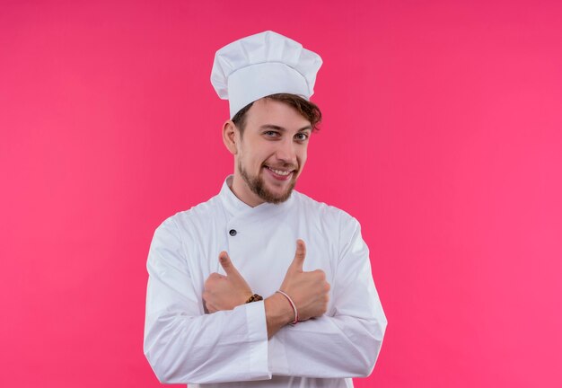 Een vrolijke jonge bebaarde chef-kok in wit uniform glimlacht en toont beide duimen terwijl hij op een roze muur kijkt