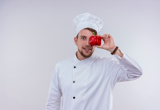 Een vrolijke jonge, bebaarde chef-kokmens die een wit fornuisuniform draagt en een hoed die rode paprika houdt en op een witte muur kijkt