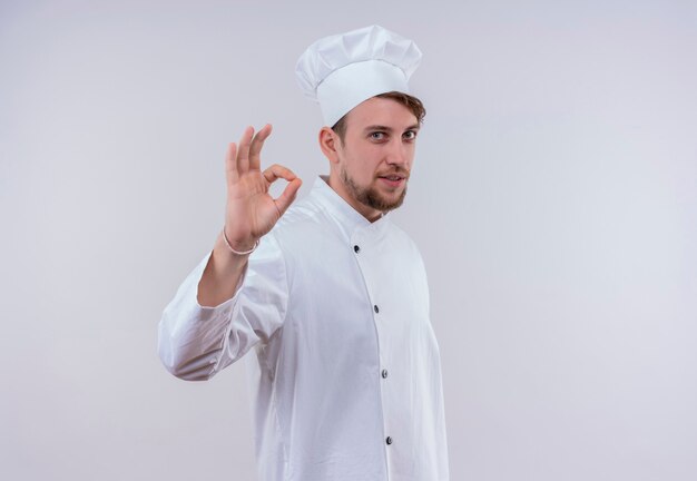 Een vrolijke jonge bebaarde chef-kokmens in wit uniform die ok gebaar op een witte muur toont