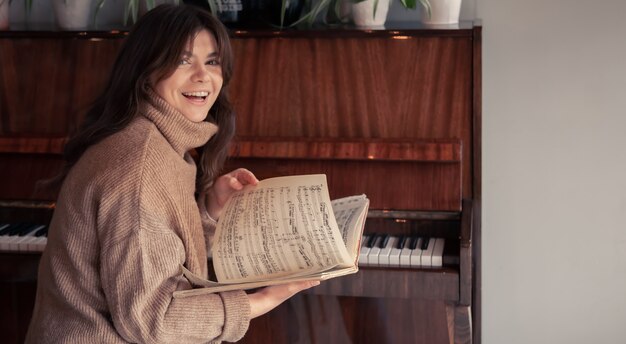 Een vrolijke jonge vrouw in een gezellige trui zit bij de piano en kijkt naar de noten.