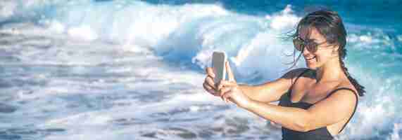 Gratis foto een vrolijke vrouw in een zwembroek maakt een selfie tegen de achtergrond van zeegolven