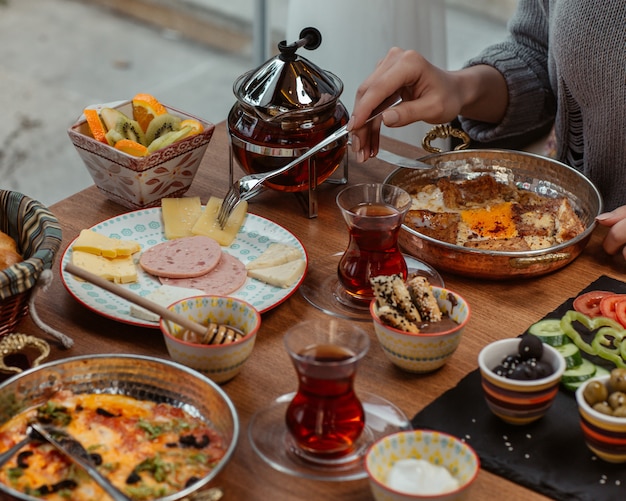 Een vrouw die ontbijtomelet eet in een pan, rond een tafel gedoneerd met olijven, kaas, salami, snoep, groenten en zwarte thee.