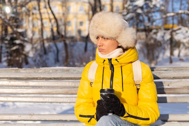 Een vrouw in de winter in warme kleren in een met sneeuw bedekt park op een zonnige dag zit op een bankje en bevriest van de kou, is ongelukkig in de winter, houdt koffie alleen