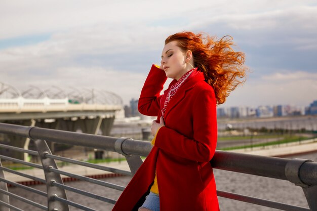 Een vrouw in een rode jas staat op de brug en trekt haar haren recht in de wind.