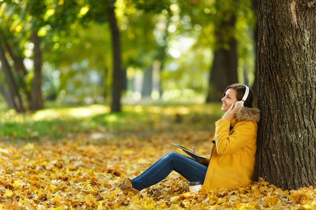 Een vrouw in gele jas en spijkerbroek zittend met een kopje koffie of thee en luisteren naar muziek onder een boom met een tablet in haar handen en koptelefoon in herfst stadspark op een warme dag. herfst gouden bladeren.