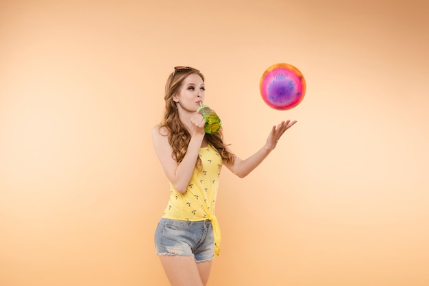 Een vrouw in lichte kleding, een geel T-shirt en een zonnebril met een spijkerbroek, houdt een bal en koude limonade in haar handen op een geïsoleerde achtergrond