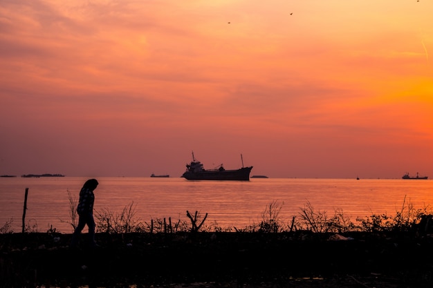 Gratis foto een vrouw lopen op de kust van de zee met een schip in het water bij zonsopgang