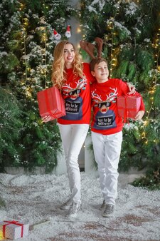 Een vrouw met mooi haar en een rode trui staat bij natuurlijke kerstbomen met een volwassen zoon en in hun handen hebben ze dozen met cadeaus