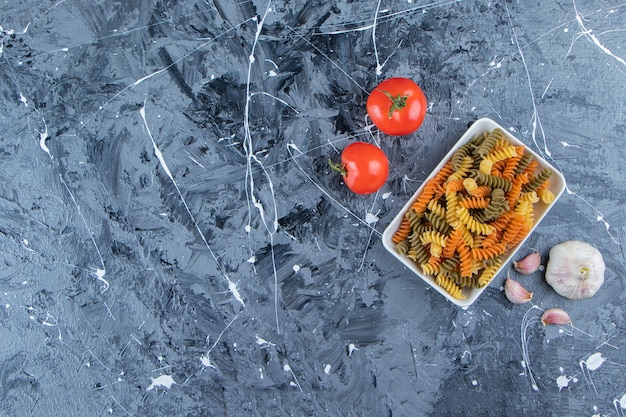 Een wit bord van multi gekleurde macaroni met verse rode tomaten en knoflook op een marmeren achtergrond.
