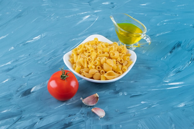 Een wit bord van rauwe pasta met olie en verse rode tomaten op een blauwe ondergrond.