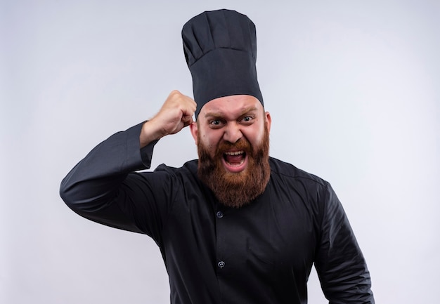 Een woedende, bebaarde chef-kok in zwart uniform die gebalde vuist opheft terwijl hij op een witte muur kijkt