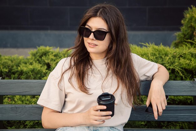 Een zaken jonge vrouw in zonnebril zit op een bankje met een kopje koffie