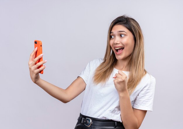 Een zeer gelukkige mooie jonge vrouw die in wit t-shirt mobiele telefoon bekijkt terwijl gebalde vuist wordt opgeheven