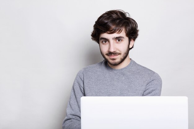 Een zelfverzekerde aantrekkelijke man met donkere baard en stijlvol kapsel zit achter een opengeklapte laptopcomputer