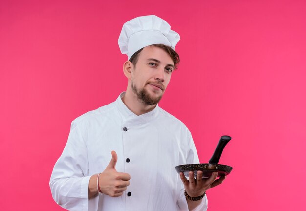 Een zelfverzekerde jonge bebaarde chef-kok in wit uniform duimen opdagen met een koekenpan in de andere hand terwijl hij op een roze muur kijkt