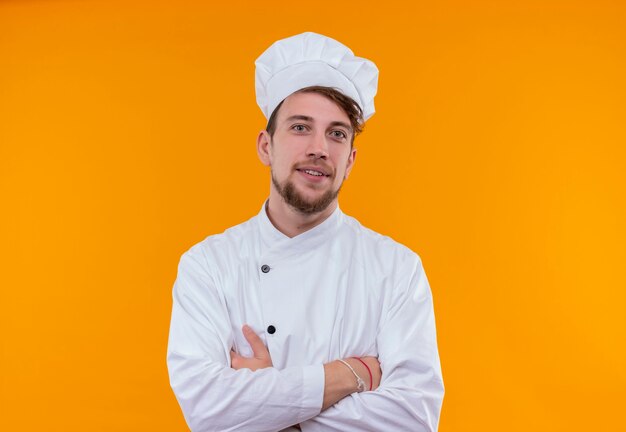 Een zelfverzekerde jonge, bebaarde chef-kokmens in wit uniform hand in hand gevouwen terwijl hij op een oranje muur keek