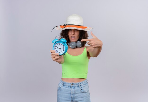 Een zelfverzekerde jonge vrouw met kort haar in een groene crop top met een zonnehoed wijzend op een blauwe wekker met wijsvinger op een witte achtergrond