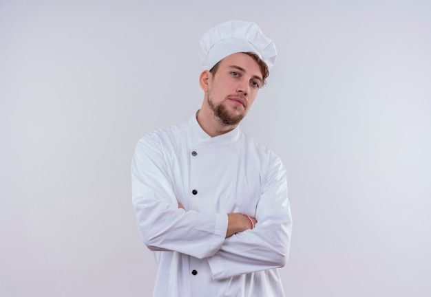 Een zelfverzekerde knappe jonge bebaarde chef-kok man gekleed in een wit fornuis uniform en hoed hand in hand gevouwen terwijl hij op een witte muur kijkt