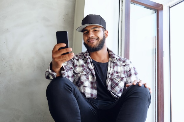 Een zwarte hipster-man gekleed in een fleece-shirt en honkbal zit in de buurt van het raam en gebruikt een smartphone.