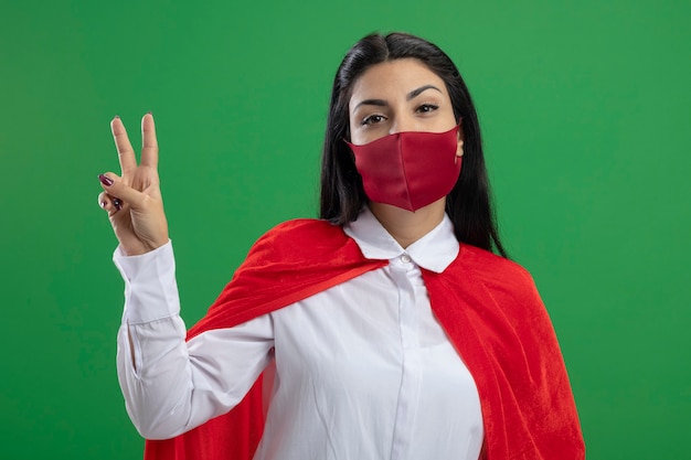 Eenvoudig jong Kaukasisch superheromeisje die masker dragen die vredesteken bekijken die camera bekijken die op groene achtergrond wordt geïsoleerd
