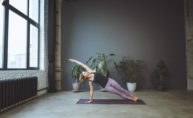 Eenzame vrouw in sportkleding doet zijplank in een ruime yogastudio Sportief fit vrouw beoefent Hatha Yoga volledige lengte grijze achtergrond Hoge kwaliteit foto