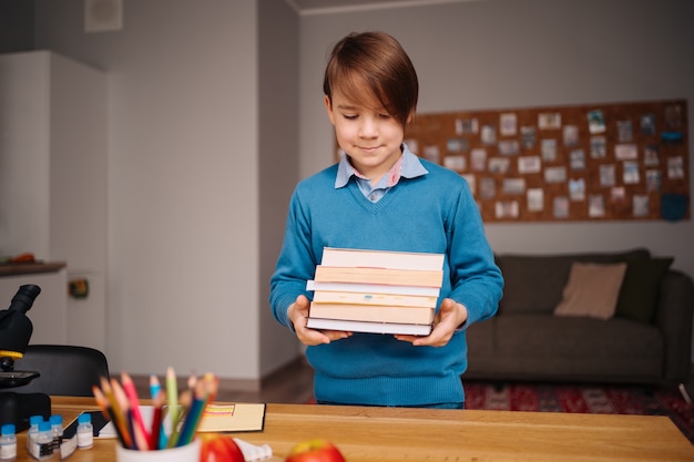 Eerste klas jongen die thuis studeert, een stapel boeken vasthoudt, zich klaarmaakt voor online les