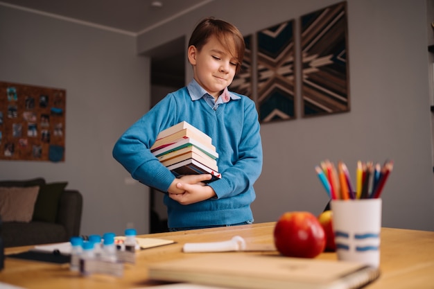 Eerste klas jongen die thuis studeert, een stapel boeken vasthoudt, zich klaarmaakt voor online les