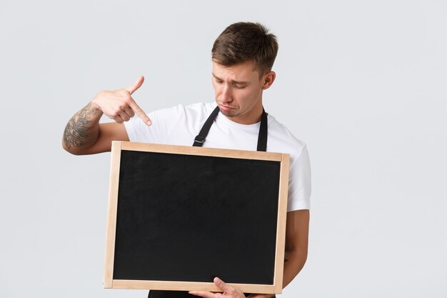 Eigenaren van kleine winkels, café- en restaurantmedewerkersconcept. Boos sombere en verontruste verkoper wijzende vinger naar krijtbord zonder tekenen, staande witte achtergrond teleurgesteld.