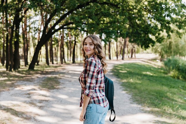 Elegant meisje met krullend kapsel over schouder kijken en genieten van wandeling in park. Goedgehumeurde dame in vrijetijdskleding die zich voordeed op de natuur.