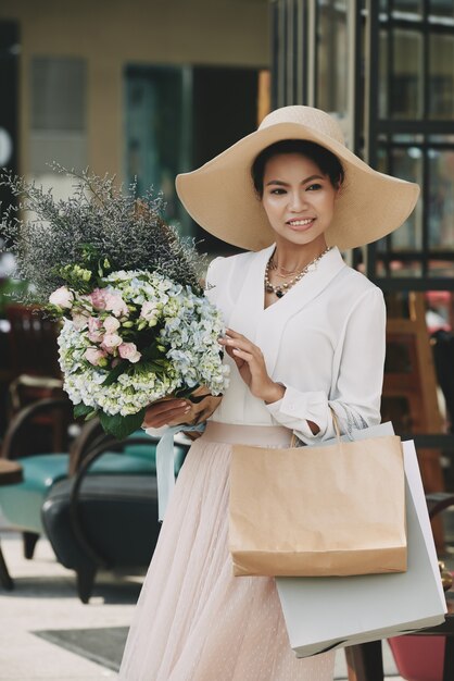 Elegante Aziatische dame in grote strohoed poseren met boodschappentassen en bloemboeket