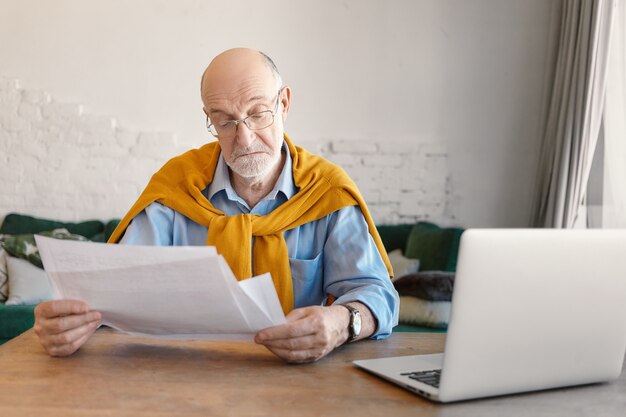 Elegante bebaarde oudere man in rechthoekige bril studeren vellen papier in zijn handen, binnenlandse financiën online thuis berekenen, met behulp van elektronische draagbaar apparaat in woonkamer interieur