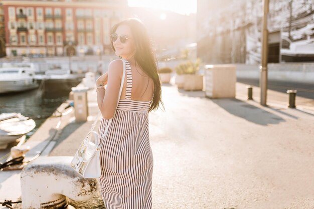 Elegante brunette vrouw in lange zomerjurk die over haar schouder kijkt en op zonnige dag naar het kanaal loopt. Outdoor Portret van gelooid meisje in zonnebril ontspannen naast kanaal in Europese stad.