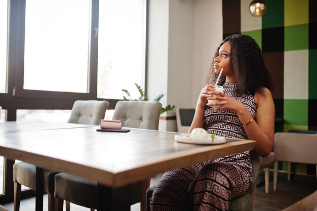 Elegante jonge Afro-Amerikaanse vrouw met lang krullend haar, gekleed in een jumpsuit die zich voordeed in café binnen met een milkshake-cocktail