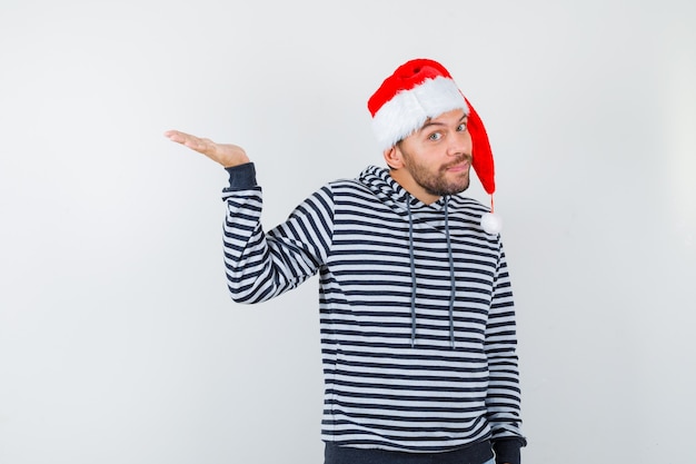 Elegante jonge man in hoodie, kerstmuts die palm opzij spreidt en aarzelend kijkt,