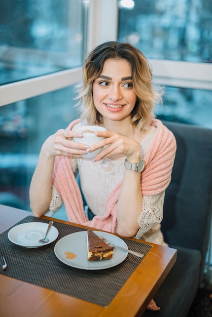 Elegante jonge positieve vrouw met een kopje drank in de buurt van smartphone en dessert aan tafel in het café