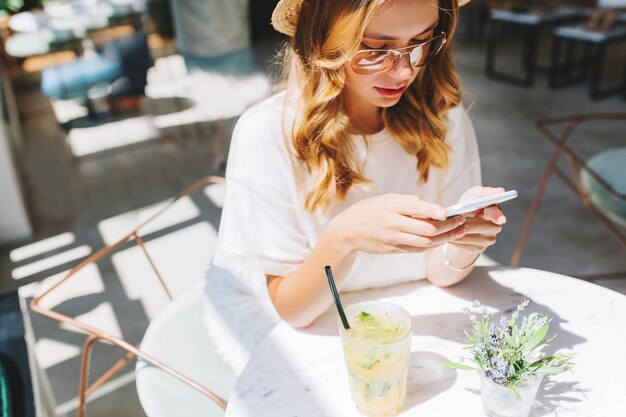 Elegante jonge vrouw in wit overhemd en trendy bril sms-bericht tijdens rust in café alleen