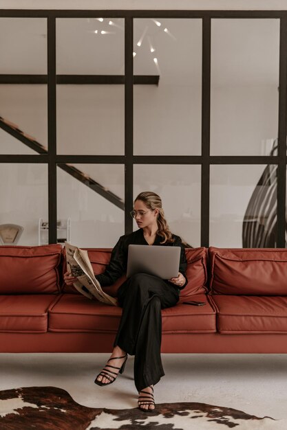 Elegante jonge vrouw in zijden zwarte jumpsuit en bril zit op een bruine bank in de woonkamer leest