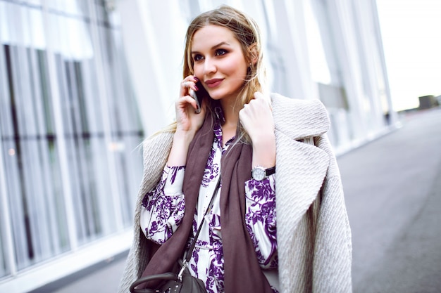 Elegante jongedame bellen op haar smartphone, luxe trendy beige jas, taupe kasjmier sjaal en gebloemde jurk dragen, poseren in de buurt van model business center.